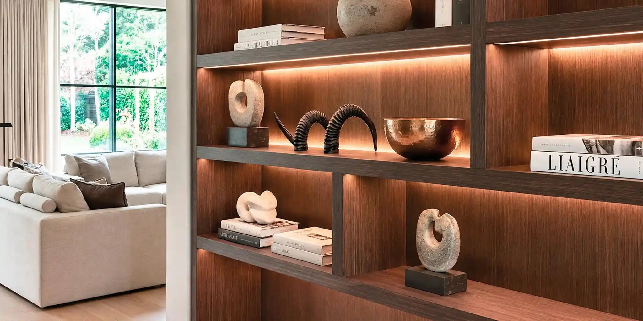 Libreria in legno illuminata a led con ripiani che espongono sculture astratte in pietra chiara, un corno decorativo scuro, una ciotola in rame e libri
