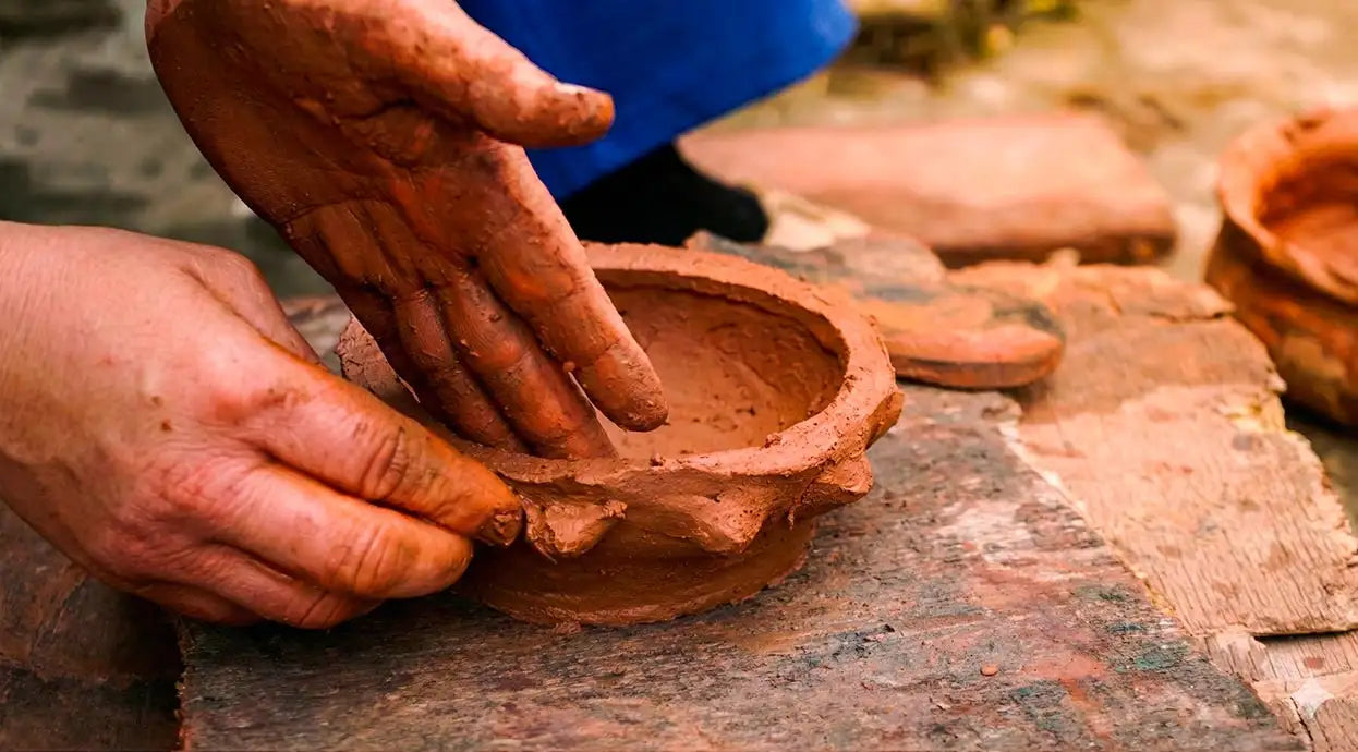 Dettaglio delle mani di un artigiana tunisina che modellano argilla rossa grezza su un tavolo da lavoro rustico, rappresentando la tradizione della ceramica fatta a mano malles per vicode
