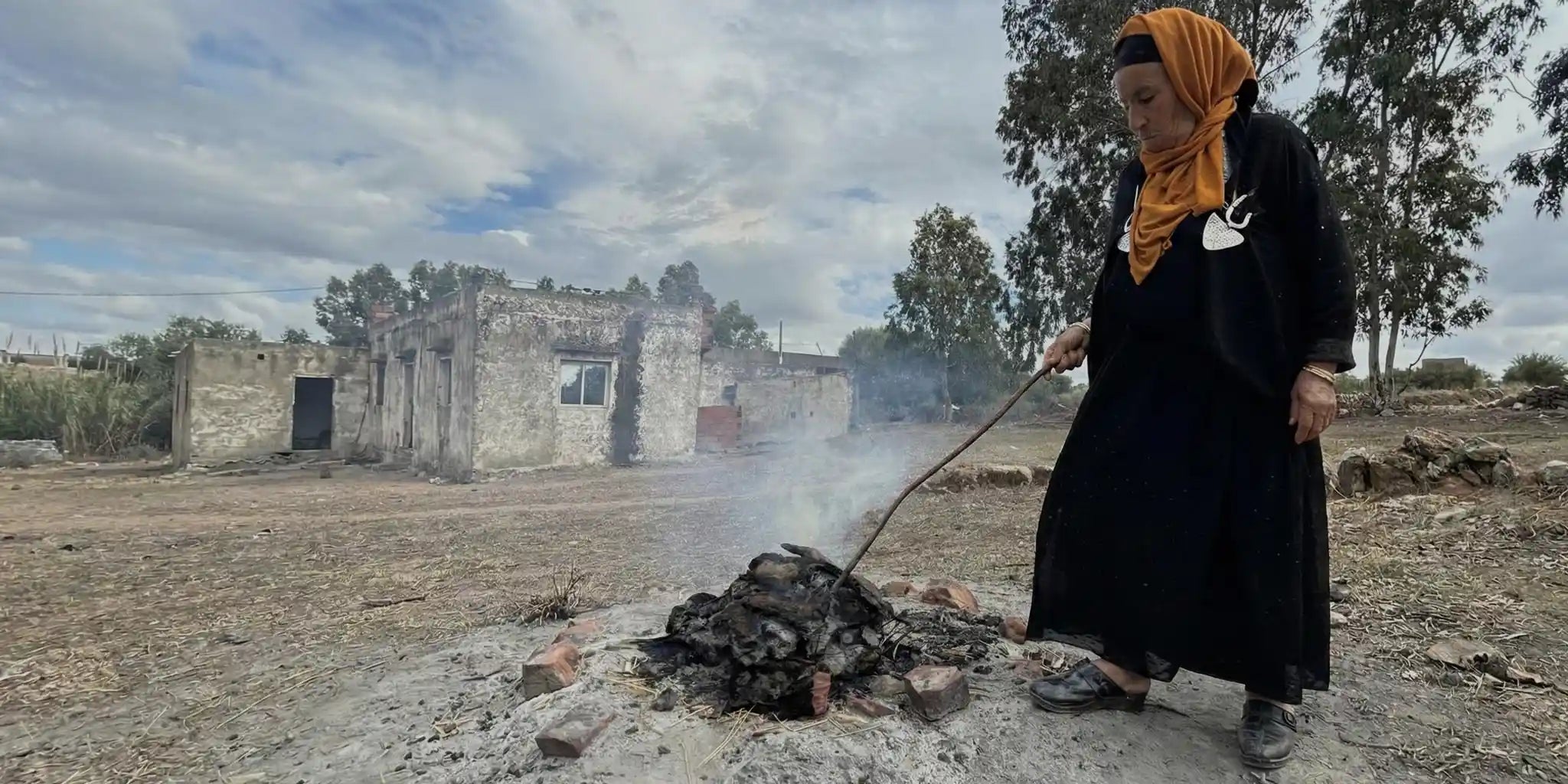 Una donna tunisina con un velo arancione utilizza un lungo bastone per alimentare un fuoco all'aperto dove vengono cotte le ceramiche artigianali Malles per vicode, in un paesaggio rurale.