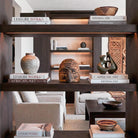 Lussuosa libreria a muro in legno scuro in un ambiente living stile Liaigre. Al centro del ripiano illuminato spicca un'antica maschera di Pulcinella in legno, circondata da pile di libri di design (tra cui 'Liaigre Works' e 'African Art'), vasi in ceramica testurizzata e sculture. Sullo sfondo, poltrone in lino e tessuti etnici.