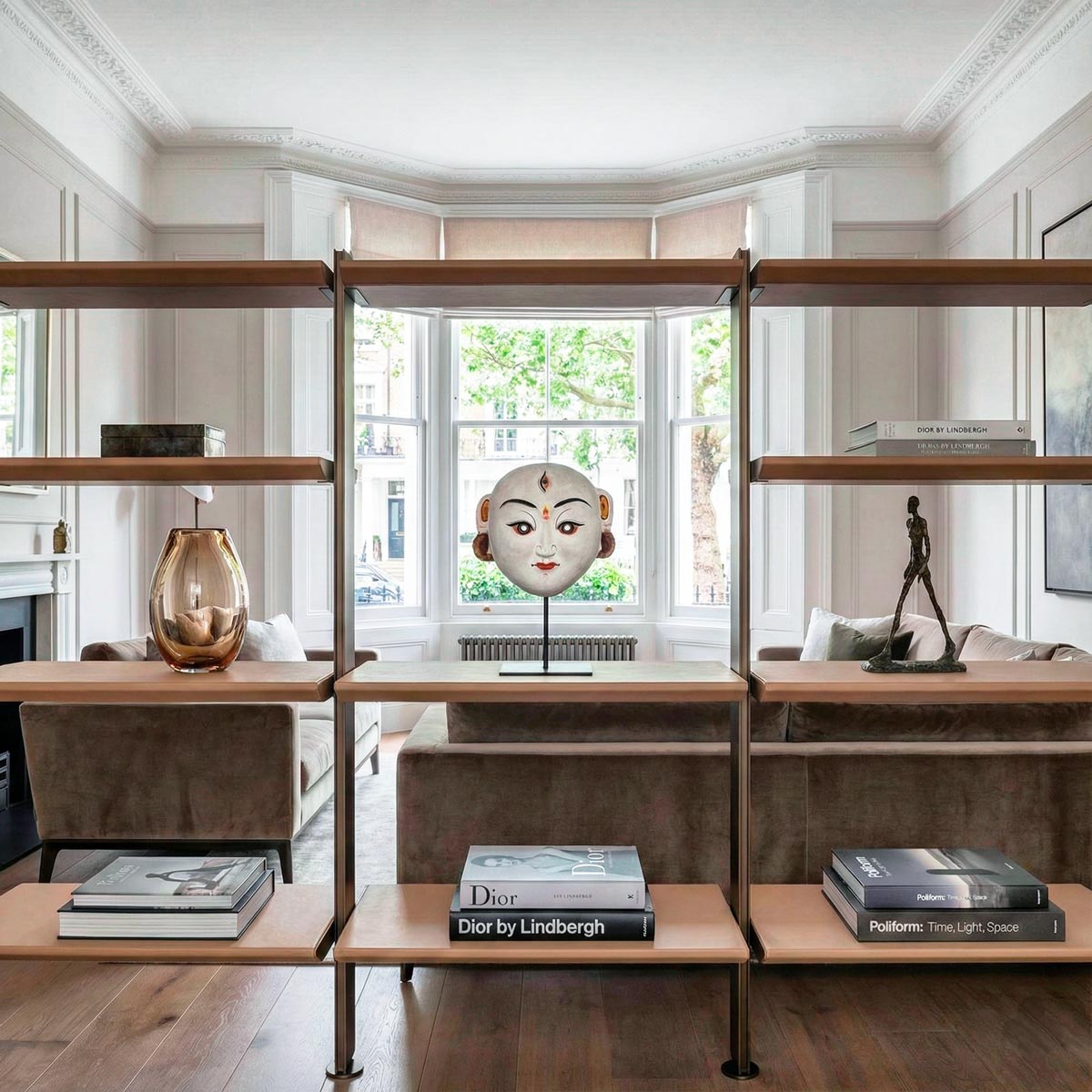 Sofisticata zona living di un appartamento londinese dallo stile contemporaneo. Su un'elegante libreria con struttura in metallo bronzato e ripiani rivestiti in pelle color biscotto, è esposta una maschera rituale di Durga in cartapesta su piedistallo, armoniosamente circondata da libri di design e oggetti d'arte in un contesto stile Poliform.