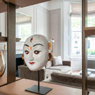 Primo piano artistico della maschera 'Ballo di Durga'. La ripresa ravvicinata evidenzia la texture artigianale della cartapesta bianca e i dettagli dipinti a mano del volto sacro. La scultura, alta 18 cm, si staglia nitida sul supporto in metallo nero, mentre lo sfondo sfocato lascia intuire un ambiente d'interni lussuoso e moderno.