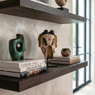 Zoom ravvicinato con prospettiva dal basso di una maschera tribale in legno intagliato e dipinto a mano, che mostra la texture invecchiata della vernice bianca, rossa e nera. La maschera è posizionata sopra i libri d'arte 'LIAIGRE 12 Projects' e 'AFRICAN ART NOW', i cui dorsi sono visibili in primo piano. Lo sfondo con la mensola e la finestra è sfocato.