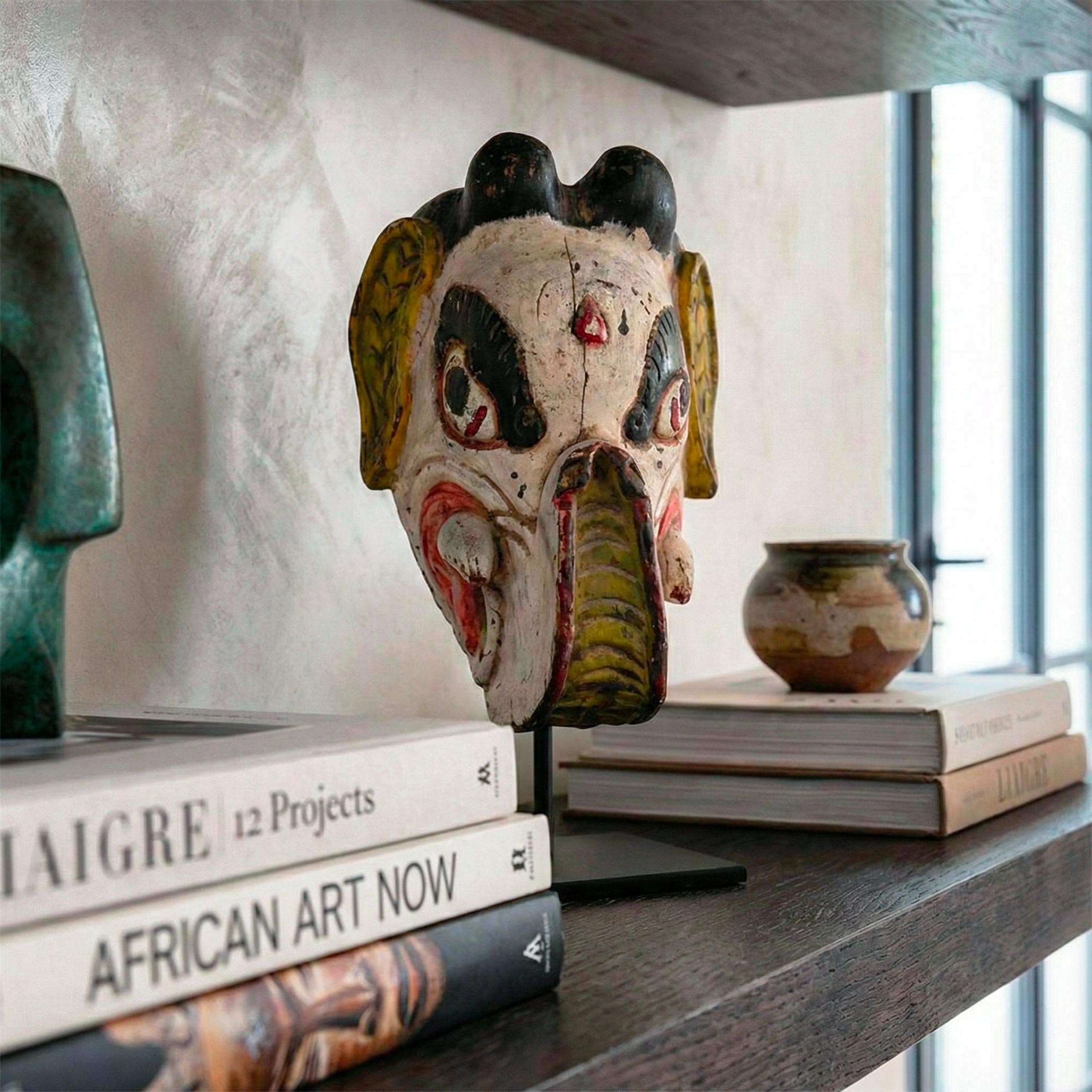 Maschera in legno policromo su piedistallo, esposta su una spessa mensola in rovere scuro in un lussuoso ambiente di design stile Liaigre. La maschera è circondata da una scultura in bronzo verde, una ciotola in ceramica e pile di libri d'arte come 'LIAIGRE 12 Projects' e 'AFRICAN ART NOW'. La luce naturale proviene da una grande finestra a tutta altezza sulla destra.
