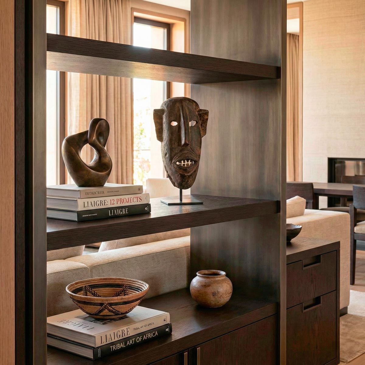 Raffinato interno in stile Liaigre che incornicia una libreria di design. Al centro, armoniosamente disposta tra volumi d'arte e oggetti preziosi, spicca una maschera africana Dogon in legno tropicale scuro alta 35 cm, creando un sofisticato contrasto tra etnico e minimalismo di lusso.