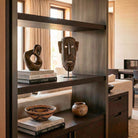 Raffinato interno in stile Liaigre che incornicia una libreria di design. Al centro, armoniosamente disposta tra volumi d'arte e oggetti preziosi, spicca una maschera africana Dogon in legno tropicale scuro alta 35 cm, creando un sofisticato contrasto tra etnico e minimalismo di lusso.