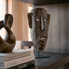 Dettaglio macro laterale della maschera tribale scolpita a mano. L'inquadratura ravvicinata mette in risalto la texture del legno invecchiato e la profonda espressività dei tratti stilizzati, come la bocca dentata. La luce morbida valorizza la patina scura della scultura staccandola dallo sfondo elegante e sfocato.