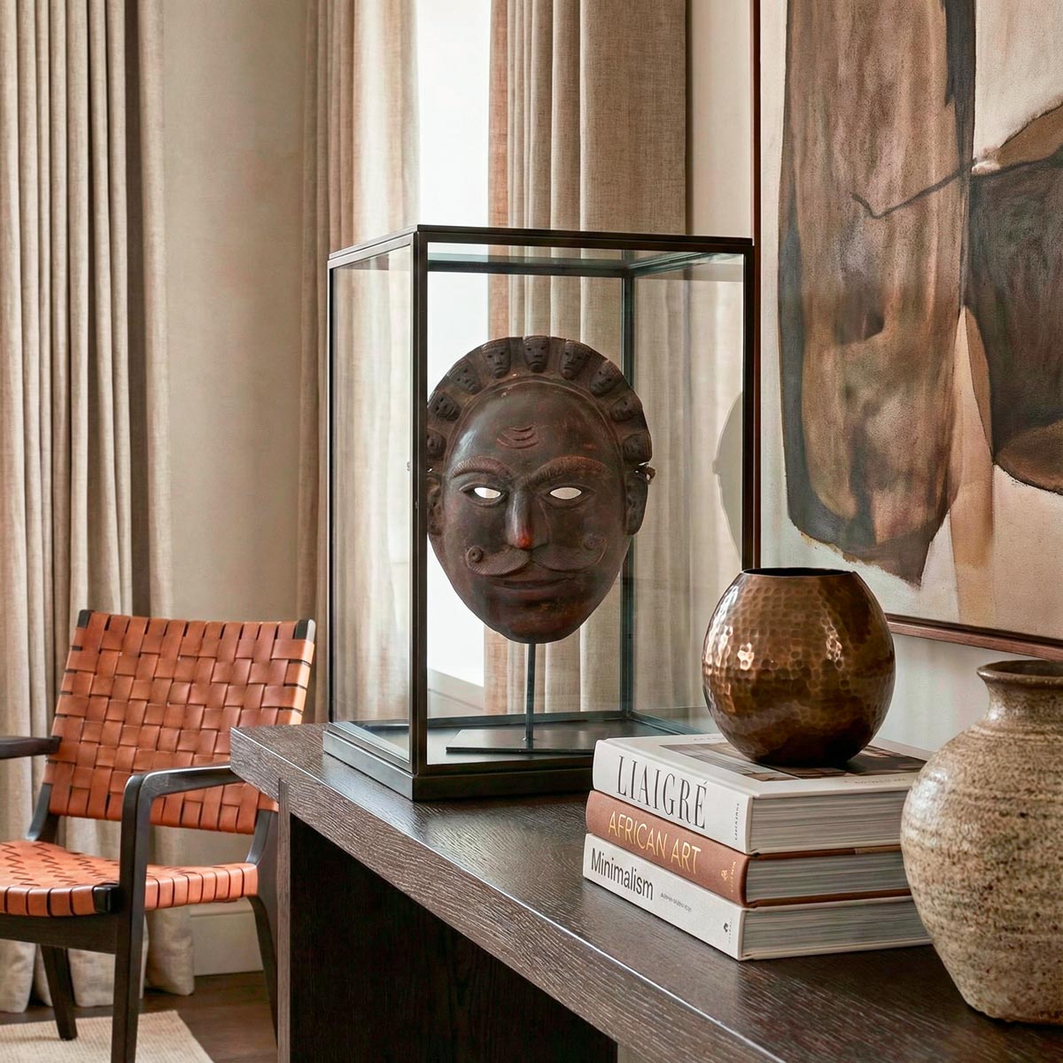 Raffinata ambientazione in stile Liaigre che espone una maschera africana Ibibio 'Re Multiforme' all'interno di una teca minimalista. La scultura in legno tropicale, alta 25 cm, è circondata da libri d'arte e oggetti preziosi, creando un perfetto equilibrio tra estetica etnica e lusso contemporaneo.