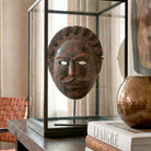 Primo piano angolato della maschera 'Re Multiforme'. La ripresa macro evidenzia la corona di piccoli volti scolpiti a mano e la patina del legno scuro. I dettagli dei baffi e dello sguardo sono a fuoco, mentre lo sfondo sfocato suggerisce un contesto di interior design esclusivo.