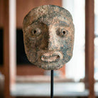 Dettaglio macro in primo piano della maschera in pietra antica. L'inquadratura ravvicinata mette in risalto la texture materica e i lineamenti del volto scolpito, sostenuto da un'asta in metallo nero, con uno sfondo neutro e sfocato che ne esalta la tridimensionalità senza distrazioni.