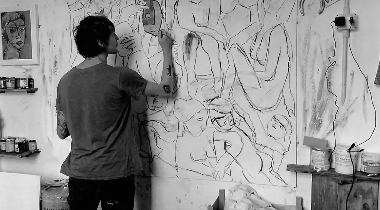 L'artista Massimiliano Precisi di spalle nel suo studio che dipinge su una grande tela piena di figure stilizzate e volti, circondato da altri quadri appesi e appoggiati.