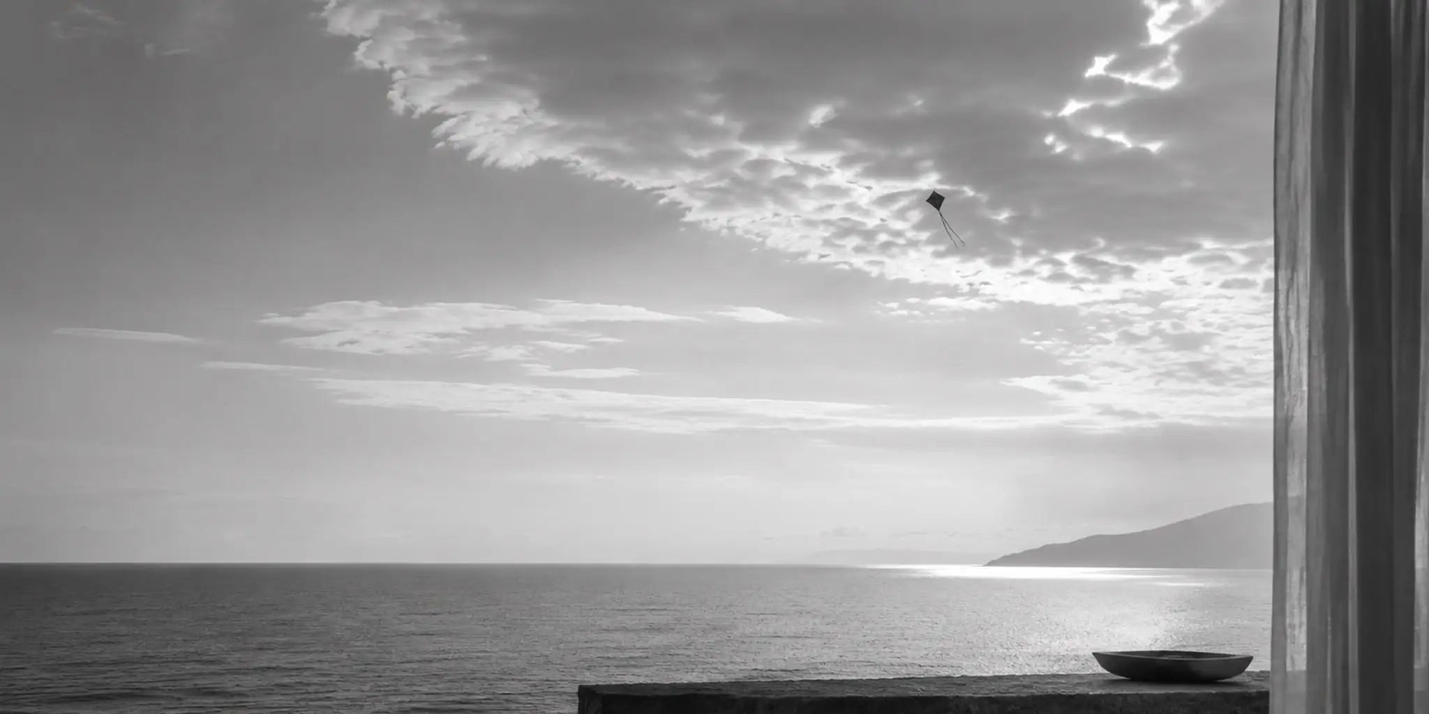 Fotografia paesaggistica in bianco e nero che ritrae un orizzonte aperto e sereno sul mare con un piccolo aquilone in volo
