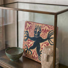 Primo piano ravvicinato all'interno di una teca espositiva in vetro e ottone. Il focus è sulla texture di una mattonella quadrata in ceramica dipinta a mano, che raffigura un diavolo nero stilizzato tra motivi rossi. In primo piano, parzialmente visibili, una ciotola in bronzo patinato e la testa di una scultura in pietra.