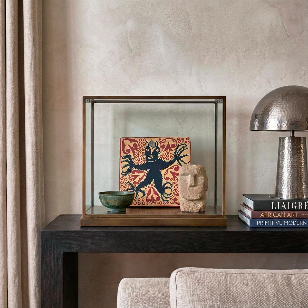 Lussuosa composizione di interior design in stile Liaigre. Su una console scura, una teca in vetro con telaio in ottone espone una mattonella in ceramica decorata con un diavolo nero su fondo rosso, affiancata da una ciotola in bronzo e una scultura Moai in pietra. A lato, una pila di libri di design (tra cui 'Liaigre' e 'African Art') e una lampada in metallo martellato.
