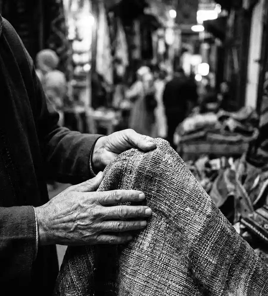 Fotografia verticale in bianco e nero che ritrae le mani vissute di un artigiano mentre toccano e mostrano un tessuto antico dalla trama ricca in un affollato mercato tradizionale (souk). Lo sfondo è sfocato, con figure umane e bancarelle, creando un'atmosfera autentica e senza tempo