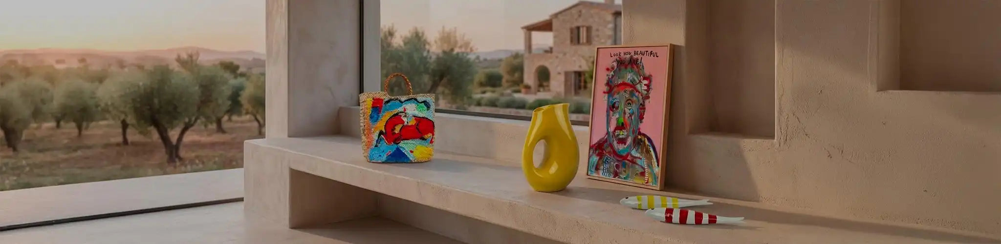 Mensola in pietra allestita con oggetti decorativi colorati: una borsa intrecciata con disegni vivaci, un vaso giallo scultoreo, una stampa incorniciata con un volto astratto e la scritta "Look how beautiful you are", e due pesciolini in ceramica a righe. Sullo sfondo, attraverso una vetrata, si vede un paesaggio di campagna con ulivi e un casale in pietra.