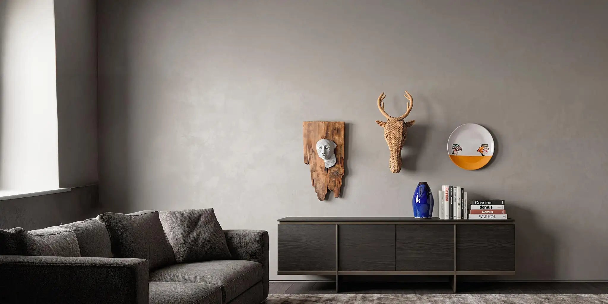 Interno di lusso in stile Christian Liaigre con elegante madia in legno scuro. Sulla parete neutra spiccano tre decorazioni artigianali: una testa di cervo in paglia intrecciata, una scultura in ceramica su legno di recupero e un piatto dipinto a mano. Sulla madia, un vaso in ceramica blu smaltata e libri di design.