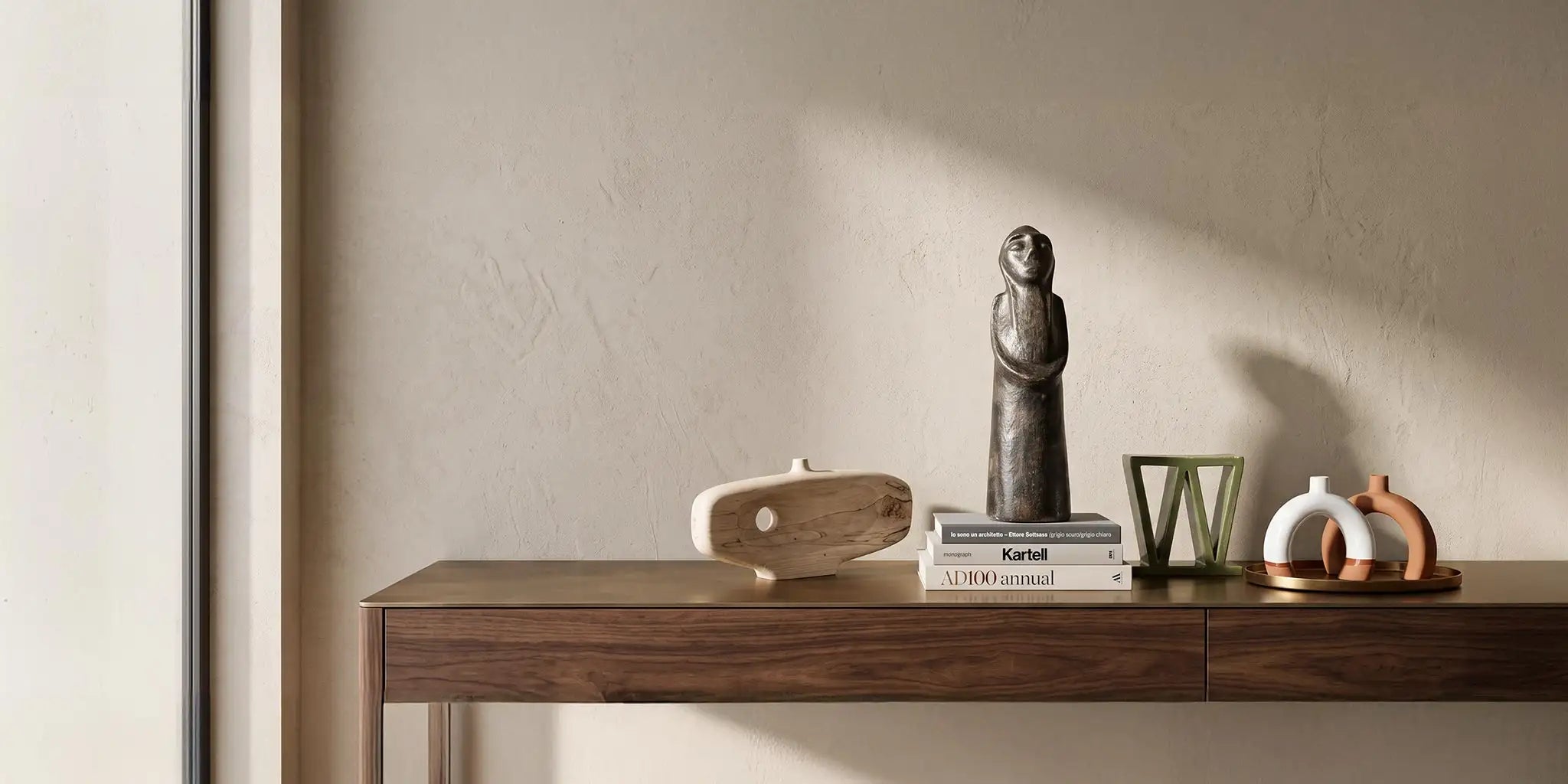Una sofisticata composizione di oggetti decorativi d'arte su una console in legno noce canaletto e piano in ottone. Include una statua femminile tunisina  su libri di design, una scultura in legno di acero,una scultura astratta inceramica smaltata e due candelabri ad arco in terracotta parzialmente smaltati, sotto una luce calda che esalta le texture.