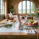 Dettaglio di una tavola rustica imbandita per un pasto festivo in una lussuosa villa toscana, con la aceteira a strisce rosse come elemento centrale tra cibo e stoviglie.