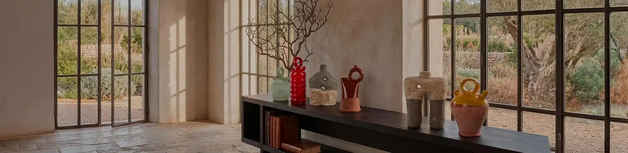 Una collezione curata di botijo in ceramica d'autore Vicode, esposta su una console in legno scuro in un ambiente architettonico in stile Liaigre. La luce naturale proveniente dalle grandi finestre illumina le diverse forme e finiture dei vasi (smaltati, materici, colorati).