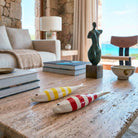 Primo piano con angolazione laterale di un tavolino da salotto in pietra grezza. Il focus è su due sculture in ceramica a forma di pesce, lunghe circa 8 cm, decorate con righe bianche e colorate (giallo e rosso). La luce naturale ne esalta la finitura lucida, mentre sullo sfondo sfocato si intravedono arredi moderni e il mare.