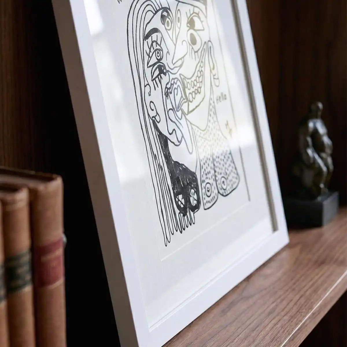 dettaglio di un quadro astratto, con disegno in bianco e nero, di un uomo e una donna che si baciano, con cornice bianca, appoggiato su una mensola in legno con libri.