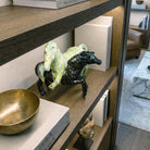 Dettaglio ravvicinato di una scultura decorativa in ceramica lucida con due cavalli in movimento. L'inquadratura dall'alto mette in risalto la finitura marmorizzata gialla e verde del primo cavallo e quella nera del secondo. L'oggetto è posizionato su una mensola in rovere illuminata, accanto a pile di libri d'arte 'Design'.
