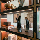 Prospettiva ravvicinata dal basso di tre sculture decorative a forma di corno in osso nero. L'illuminazione integrata nello scaffale in legno scuro esalta la texture rigata e lucida dei corni. A sinistra sono visibili libri di design 'Liaigre' impilati e una scatola in legno, mentre lo sfondo del salotto rimane sfocato per dare profondità.