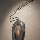 Dettaglio di un'opera d'arte contemporanea in ferro battuto installata su una parete a calce. La scultura è caratterizzata da una notevole profondità materica e bordi spessi. L'illuminazione drammatica mette in risalto la texture ruvida del metallo e il piccolo inserto pittorico centrale.