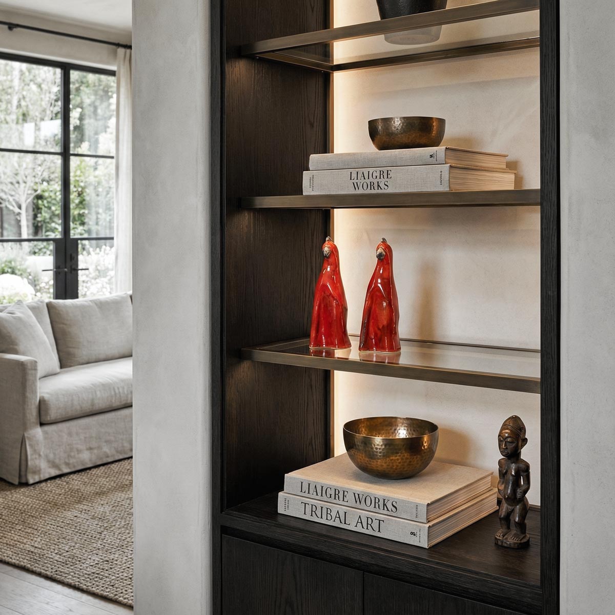 Coppia di sculture artigianali in ceramica rossa a forma di pappagallo, esposte come accento di colore su una mensola in vetro e bronzo illuminata. La libreria a muro in legno scuro stile Liaigre contiene anche libri d'arte ('Liaigre Works', 'Tribal Art'), ciotole in bronzo e una statuetta etnica, in un salotto luminoso e sofisticato