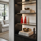 Coppia di sculture artigianali in ceramica rossa a forma di pappagallo, esposte come accento di colore su una mensola in vetro e bronzo illuminata. La libreria a muro in legno scuro stile Liaigre contiene anche libri d'arte ('Liaigre Works', 'Tribal Art'), ciotole in bronzo e una statuetta etnica, in un salotto luminoso e sofisticato