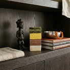 Primo piano di una composizione di design con quattro mattoncini di ceramica smaltata impilati, ognuno con il nome di una stagione in rilievo: PRIMAVERA (verde), ESTATE (giallo), AUTUNNO (marrone) e INVERNO (bianco). A sinistra, una scultura africana in legno scuro raffigurante una figura femminile. Sullo sfondo, una ciotola in metallo martellato con una collana di perle ambrate e dei libri, il tutto su un ripiano di boiserie in rovere scuro in un ambiente di lusso in stile Liaigre.