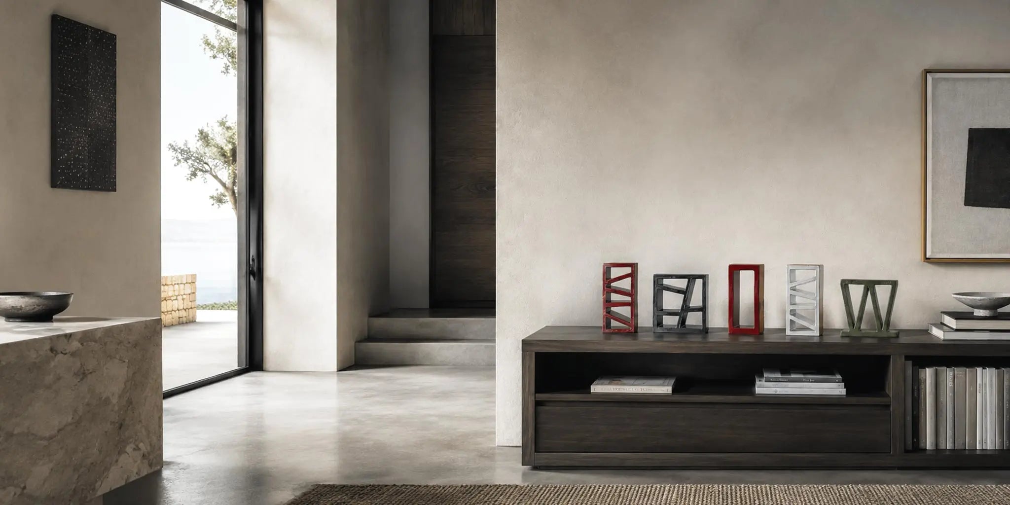 Sculture geometriche in ceramica smaltata disposte su una madia in legno scuro, in un elegante living mediterraneo con pareti materiche e vista sul mare.