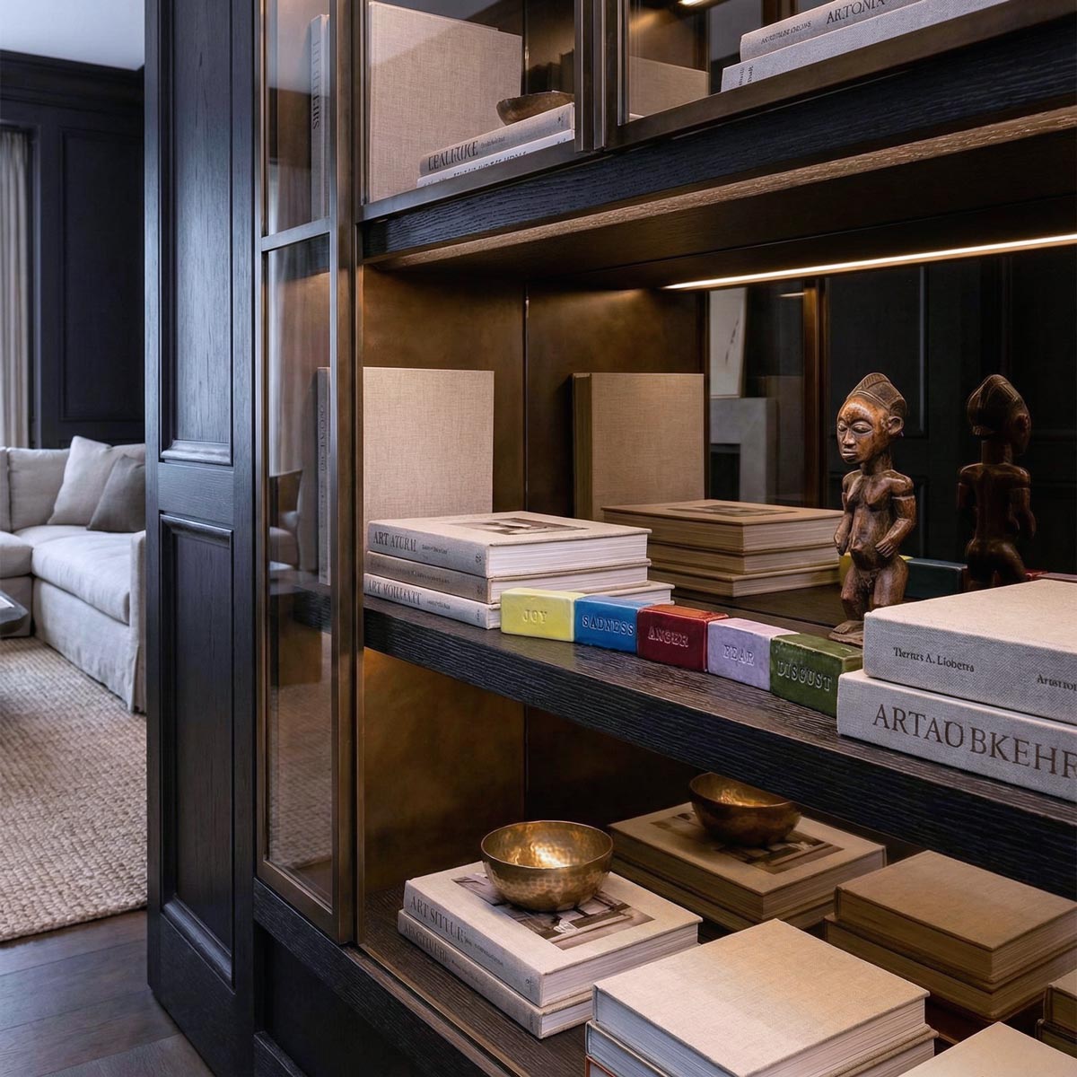 Set di cinque mattoncini in ceramica smaltata colorata con incisi i nomi delle emozioni (JOY, SADNESS, ANGER, FEAR, DISGUST), esposti in fila su una mensola illuminata in una lussuosa teca in legno scuro e bronzo. La composizione include libri d'arte rilegati in lino, una statuetta etnica in legno e una ciotola in bronzo in un salotto elegante.