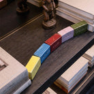 Dettaglio ravvicinato dall'alto dei mattoncini in ceramica colorata 'Emozioni'. L'inquadratura mette in risalto la finitura lucida dello smalto e le lettere incise sui blocchi (giallo, blu, rosso, rosa, verde), disposti in una leggera curva su un ripiano in legno scuro con venature visibili, circondati da pile di libri