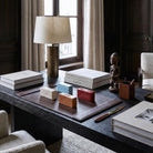 Set di quattro mattoncini in ceramica smaltata con i nomi degli elementi (ARIA, ACQUA, TERRA, FUOCO), disposti su un sottomano in pelle marrone su una scrivania direzionale in legno scuro. L'ambiente lussuoso include una lampada in ottone, libri di design e una scultura africana in legno vicino a una finestra.