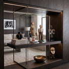 Dettaglio ravvicinato dall'alto delle due sculture in cartapesta raffiguranti un uomo e una donna. L'inquadratura mette in risalto la texture ruvida e i colori dipinti a mano dei personaggi, posizionati su una mensola in legno scuro all'interno di una teca di vetro illuminata a LED. A lato sono visibili libri d'arte e un vaso in ceramica nera.