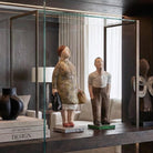 Coppia di sculture figurative in cartapesta dipinta a mano, esposte come pezzi centrali all'interno di una lussuosa teca in vetro e bronzo con illuminazione integrata. La libreria in legno scuro a tutta parete contiene anche libri di design, un vaso scultoreo e manufatti etnici in legno e metallo, in un salotto elegante.