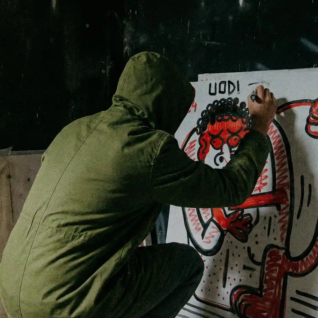Uno scatto rubato in un ambiente urbano decadente e materico, che cattura l'artista Uodi nell'atto di completare un'opera che fonde street art e pittura su tela.