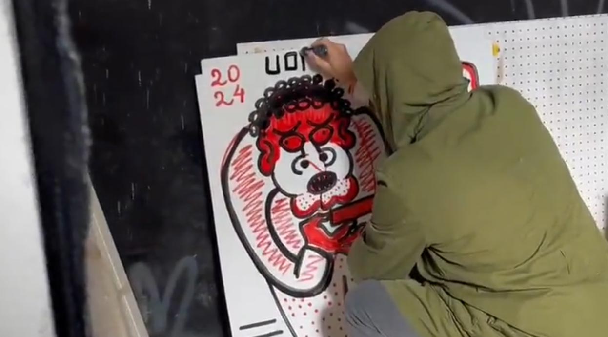 L'artista Uodi, con indosso una giacca verde, è accovacciato mentre dipinge a mano libera una grande pantera rossa stilizzata con contorni neri spessi su una tela bianca con griglie, firmata "UODI" in alto per la collezione vicode