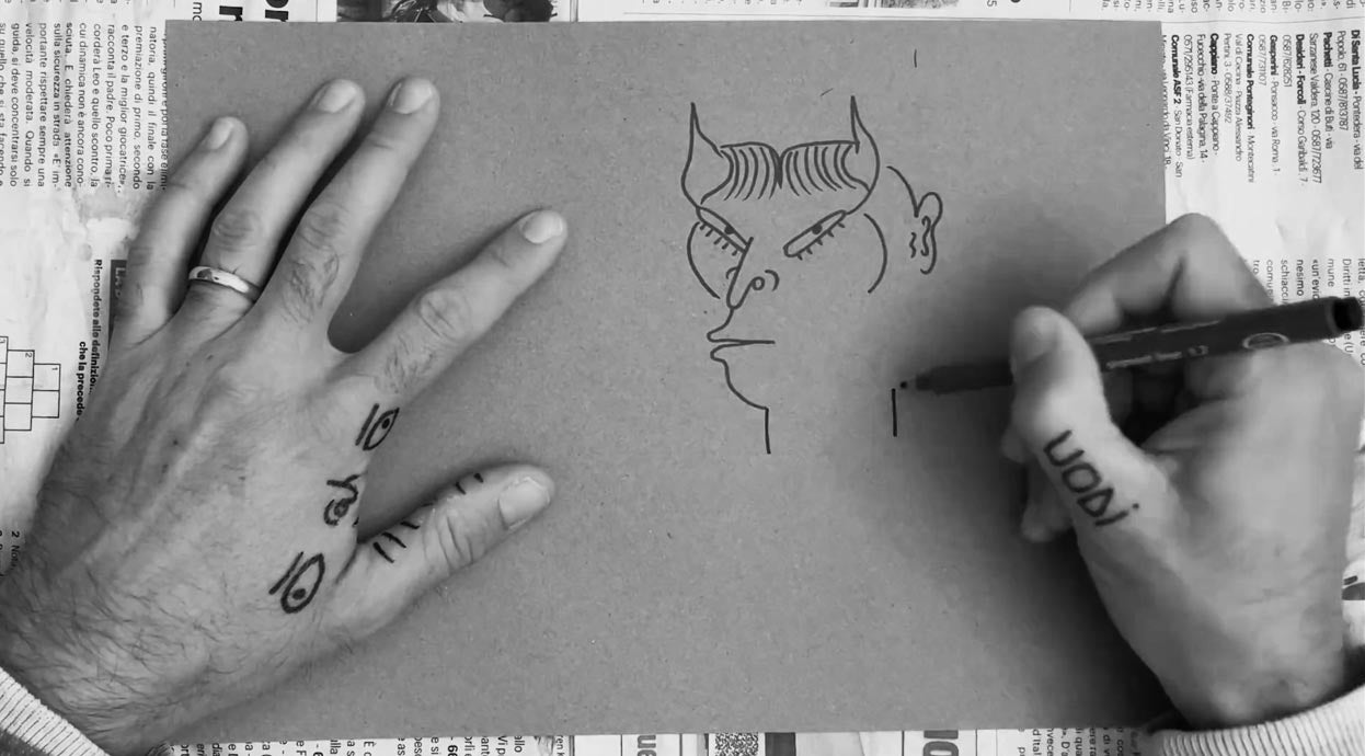 Primo piano delle mani tatuate di Uodi che, con un pennarello nero, tracciano su un foglio di carta grezza il profilo di un personaggio fantastico con corna e grandi occhi, pezzo unico della collezione uodi x vicode