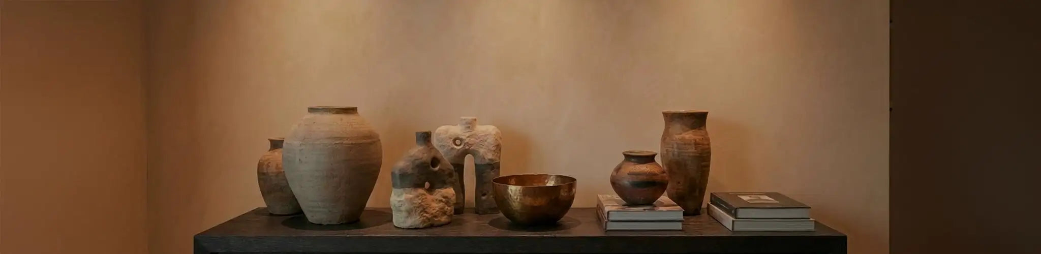 Una selezione curata di vasi artigianali in ceramica e sculture materiche dai toni naturali, disposti su una console in legno scuro. L'illuminazione d'accento calda esalta le texture contro una parete beige lavorata a calce.
