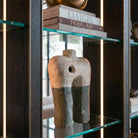 Primo piano con prospettiva leggermente dal basso della scultura astratta in cartapesta bicolore. L'inquadratura ravvicinata enfatizza la texture materica e grezza della superficie beige superiore e della base grigio scuro. La scultura poggia su un ripiano in vetro, affiancata dalla struttura verticale in legno e dalla striscia LED accesa, con i dorsi dei libri 'Liaigre' e 'Tribal Art' visibili nel ripiano superiore.