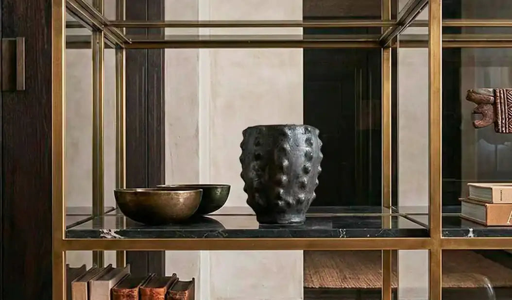 Vaso scultoreo nero Malles x Vicode, interamente fatto a mano e cotto nel fuoco in Tunisia. La superficie materica e irregolare del vaso, esposto su un'elegante libreria in vetro e ottone, esalta il contrasto tra design contemporaneo e antiche tecniche artigianali.