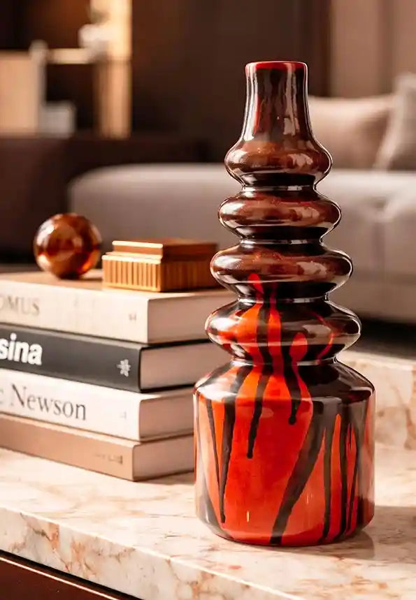 Vaso di design Vicode in ceramica con silhouette a balze e finitura lucida effetto colato nei colori marrone cioccolato e rosso vibrante, appoggiato su un tavolino in marmo accanto a libri d'arte.