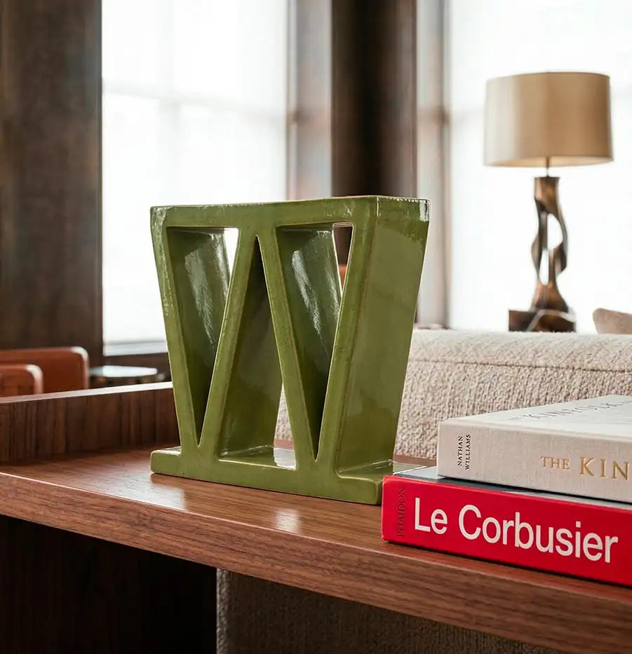 Elegante scultura Vicode in ceramica verde a forma di W esposta su una mensola in legno accanto a un libro di Le Corbusier, ideale come elemento d'arredo per progetti di architetti nei settori contract e hotellerie.